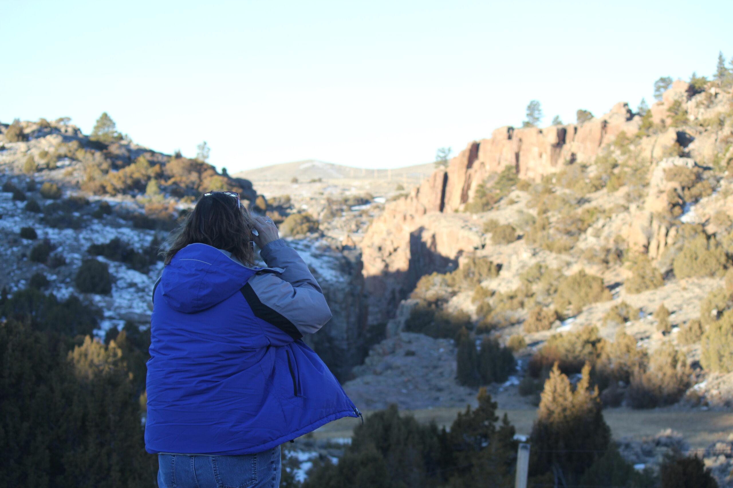 A woman in a blue jacket stands over a valley with binoculars in her hand as she works on a Wyoming Bureau of Land Management bald eagle survey. 
